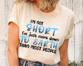 May include: Cream-colored t-shirt with the text "I'm not short, I'm just more down to earth than most people" in blue and black lettering. The shirt has rolled-up sleeves and is paired with blue jeans.
