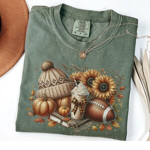 May include: Green t-shirt with a fall-themed design featuring a brown football, a pumpkin, a book, a cup of coffee with whipped cream, a sunflower, and a brown knit hat with a white pom-pom.