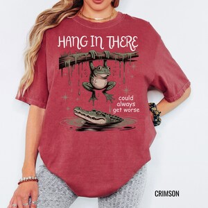May include: Crimson-colored t-shirt with the phrase "Hang in There" above a cartoon frog hanging from a branch, with an alligator below. The text "could always get worse" is also included. The shirt is a relaxed fit.