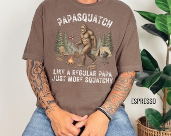 May include: Espresso-colored t-shirt with the text "Papasquatch" and a graphic of Bigfoot at a campsite. The graphic includes a campfire, tent, and trees. The text "Like a regular papa, just more squatchy" is also printed on the shirt.