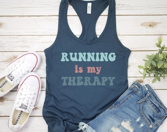 May include: A navy blue tank top with the text "RUNNING is my THERAPY" in a retro font with pastel blue, green, and pink colors. The tank top is laying on a white wood surface with a pair of white canvas shoes and a pair of blue denim shorts.