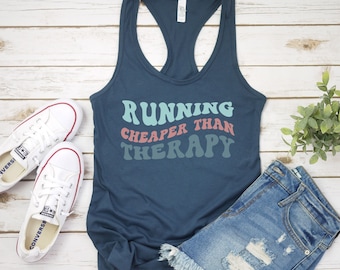 May include: A navy blue tank top with the text "RUNNING CHEAPER THAN THERAPY" in a retro font with colorful accents. The text is in a light blue, pink, and green color scheme.