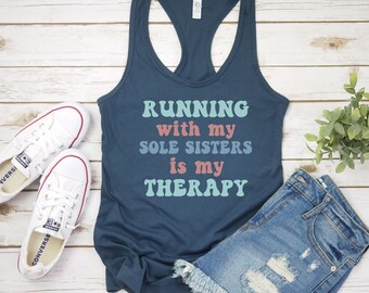 May include: A navy blue tank top with the text "RUNNING with my SOLE SISTERS is my THERAPY" in a retro font with pink, blue, and green colors. The tank top is paired with denim shorts and white sneakers.