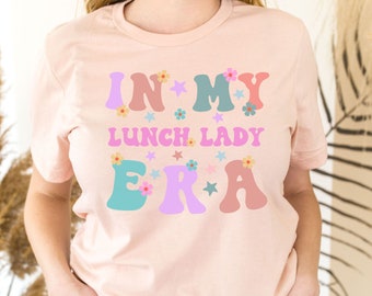 May include: A pink t-shirt with the text "In My Lunch Lady Era" in colorful, retro-style lettering with flowers and stars.