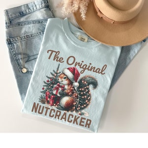 May include: A light blue t-shirt with a brown squirrel wearing a Santa hat and holding a red gift box. The squirrel is surrounded by a Christmas tree and lights. The text "The Original Nutcracker" is printed on the shirt.
