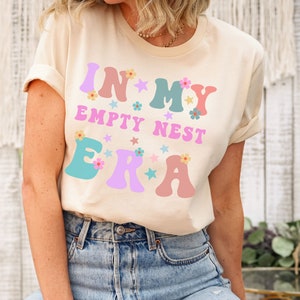 May include: A light beige t-shirt with the text "In My Empty Nest Era" in colorful, stylized letters with flowers and stars.