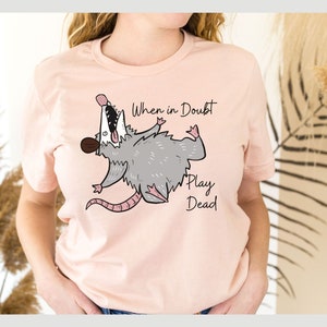 Pu&ograve; includere: Una maglietta rosa con un opossum dei cartoni animati che finge di essere morto. Il testo "When in doubt, play dead" &egrave; stampato sulla maglietta.