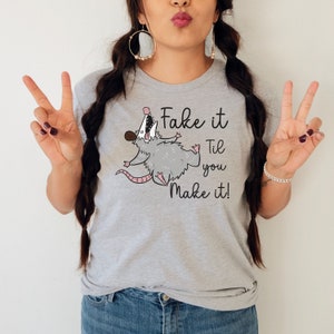 Puede incluir: Camiseta gris con un opossum de dibujos animados y el texto "Fake it Til you Make it!"