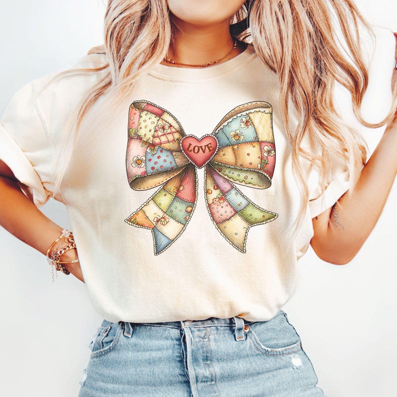 May include: Cream-colored t-shirt featuring a large, colorful bow design. The bow is made of patchwork fabric in various pastel colors, with a red heart in the center that reads "LOVE". The bow is decorated with small flowers.