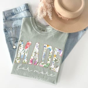 May include: Sage green t-shirt with the word "MADE" in large floral letters and the words "for more" in cursive. The shirt is folded next to a pair of light wash jeans, a tan hat, and a gold necklace.
