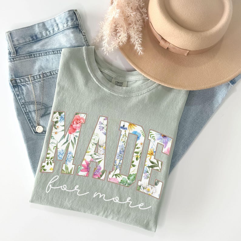 May include: Sage green t-shirt with the word "MADE" in large floral letters and the words "for more" in cursive. The shirt is folded next to a pair of light wash jeans, a tan hat, and a gold necklace.
