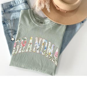 May include: A light green t-shirt with a floral print design that spells out the word "Grandma".