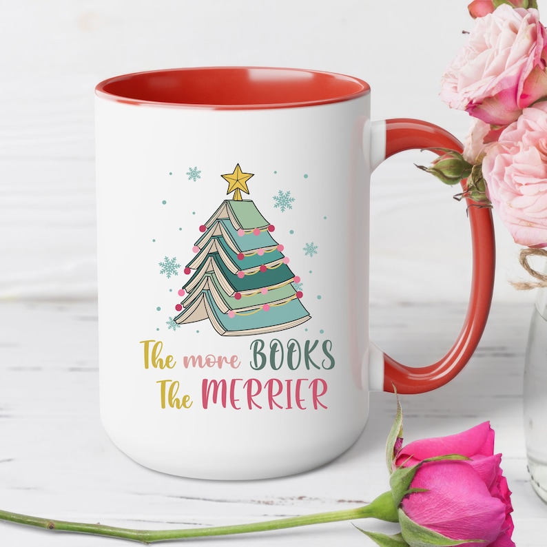 May include: White ceramic mug with a red rim and handle. The mug features a Christmas tree made of stacked books with a star on top. The text "The more books the merrier" is printed below the tree.