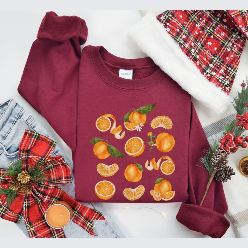 May include: A maroon sweatshirt with a graphic print of oranges and orange peels. The sweatshirt has a crew neck and long sleeves.