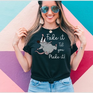 Puede incluir: Camiseta negra con un opossum de dibujos animados y el texto "Fake it Til you Make it!"