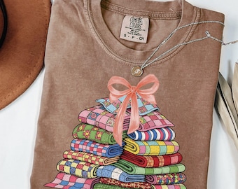 May include: A brown Comfort Colors t-shirt with a graphic of a stack of colorful quilts tied with a pink bow. A silver necklace with a small pendant is draped on the shirt. The shirt is folded and displayed on a white surface.