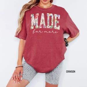 May include: Crimson-colored t-shirt with the words "MADE" and "for more" in a floral design. The shirt has short sleeves and a crew neck. The word "CRIMSON" is printed in the bottom right corner.