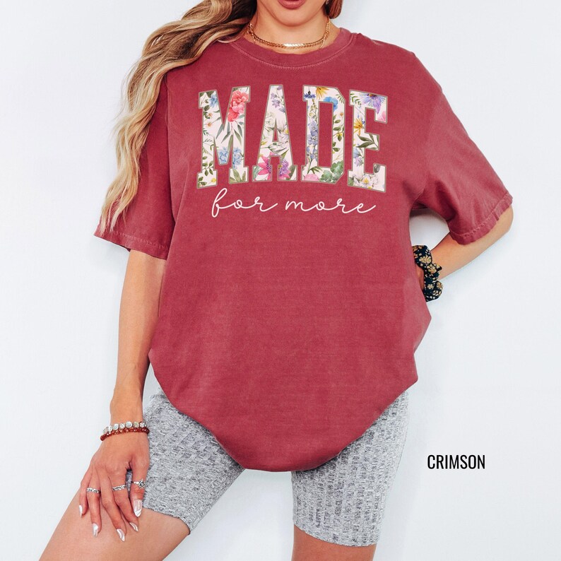 May include: Crimson-colored t-shirt with the words "MADE" and "for more" in a floral design. The shirt has short sleeves and a crew neck. The word "CRIMSON" is printed in the bottom right corner.