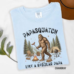 May include: Pale blue t-shirt featuring a Bigfoot illustration with a sausage over a campfire. Text reads "PAPASQUATCH LIKE A REGULAR PAPA CHAMBRAY". Includes trees, a tent, and a barbecue.