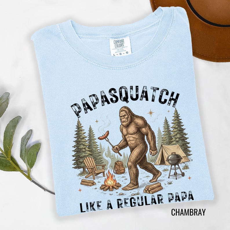 May include: Pale blue t-shirt featuring a Bigfoot illustration with a sausage over a campfire. Text reads "PAPASQUATCH LIKE A REGULAR PAPA CHAMBRAY". Includes trees, a tent, and a barbecue.
