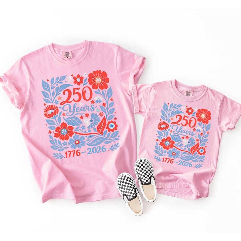 May include: Two pink t-shirts, one adult and one child size, with a floral design and the text "250 Years 1776-2026". Two pairs of black and white checkered slip-on shoes are in the foreground.