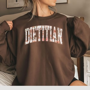 May include: A brown sweatshirt with the word "DIETITIAN" in large, floral-patterned letters across the chest. The floral pattern includes pink and white flowers. The sweatshirt has a crew neck and long sleeves.