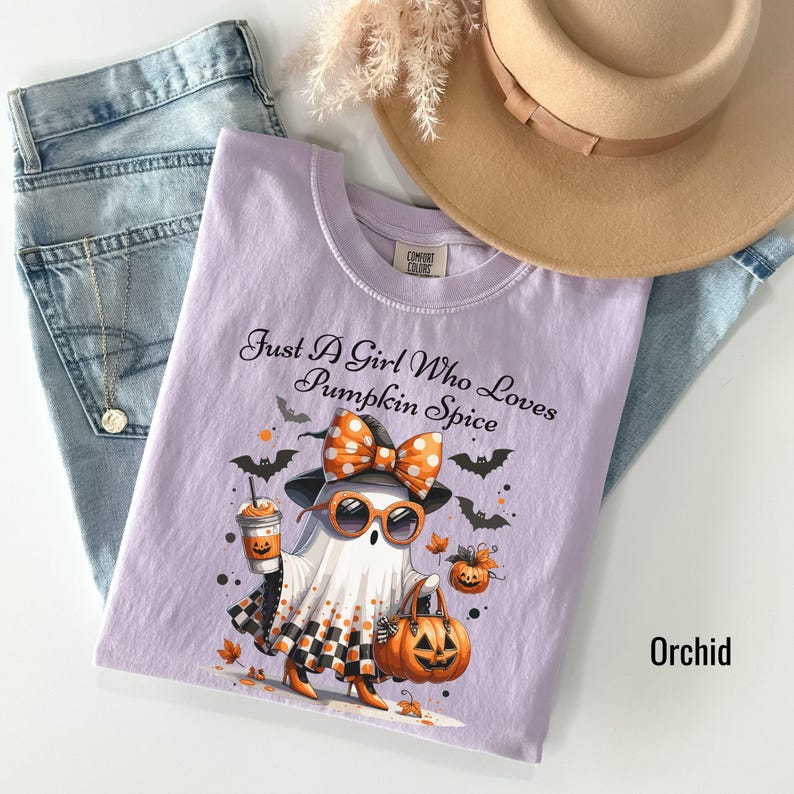 May include: Orchid-colored t-shirt with the text "Just A Girl Who Loves Pumpkin Spice" and a graphic of a ghost wearing sunglasses, a bow, and holding a pumpkin and coffee. The shirt is paired with blue jeans and a tan hat.