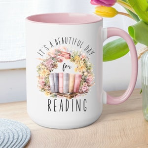 May include: White ceramic mug with a pink interior and handle. The mug features a floral wreath design with books and the text "It's a beautiful day for reading" in black lettering. The mug is a standard size.