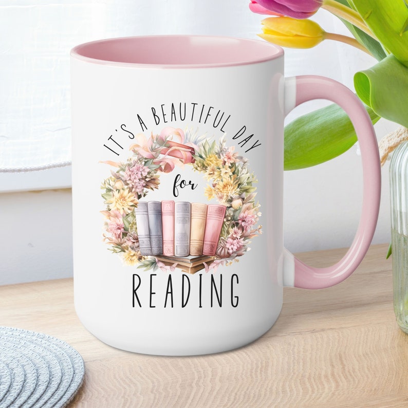 May include: White ceramic mug with a pink interior and handle. The mug features a floral wreath design with books and the text "It's a beautiful day for reading" in black lettering. The mug is a standard size.