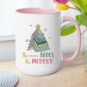 May include: White ceramic mug with a pink rim and handle. The mug features a Christmas tree made of stacked books with a star on top. The text "The more books the merrier" is printed below the tree.