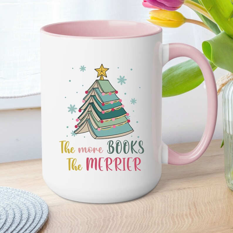 May include: White ceramic mug with a pink rim and handle. The mug features a Christmas tree made of stacked books with a star on top. The text "The more books the merrier" is printed below the tree.