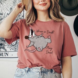 Pu&ograve; includere: Una maglietta rosa con un opossum dei cartoni animati che finge di essere morto e il testo "When in Doubt Play Dead".