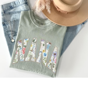 May include: Sage green t-shirt with the word "NANA" in a floral design. The shirt is paired with light wash denim jeans, a tan hat, and a gold necklace. A sprig of dried flowers adds a decorative touch.