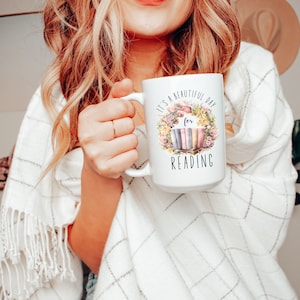May include: White ceramic mug with the text "It's a beautiful day for READING" surrounded by a floral wreath and a stack of books. The mug has a large handle and is held by a person.