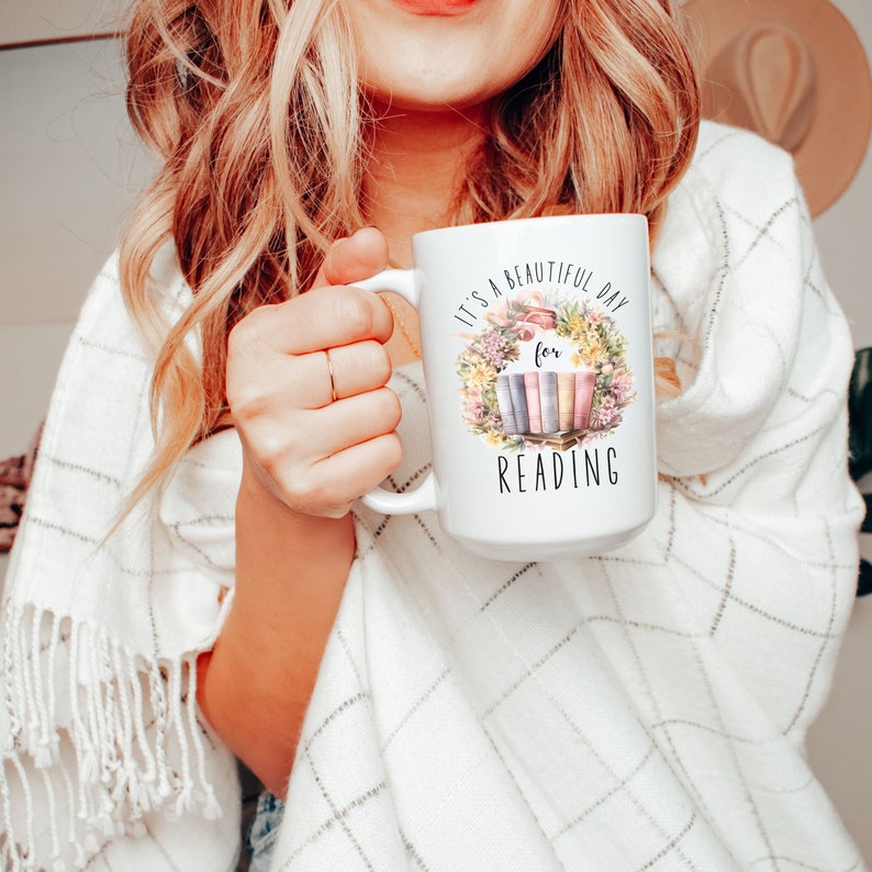 May include: White ceramic mug with the text "It's a beautiful day for READING" surrounded by a floral wreath and a stack of books. The mug has a large handle and is held by a person.
