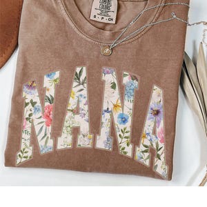 May include: A brown Comfort Colors t-shirt with the word "NANA" in large, floral-patterned letters. A silver necklace with a pendant is draped over the shirt. The shirt is a small size.