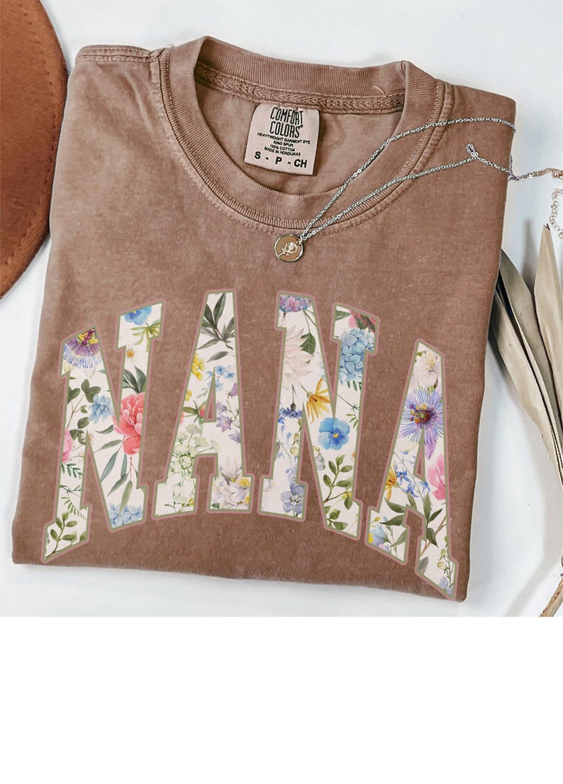 May include: A brown Comfort Colors t-shirt with the word "NANA" in large, floral-patterned letters. A silver necklace with a pendant is draped over the shirt. The shirt is a small size.