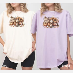 May include: Two t-shirts with a fall-themed design featuring a coffee cup, a football, pumpkins, and a hat. The t-shirts are in ivory and orchid colors.