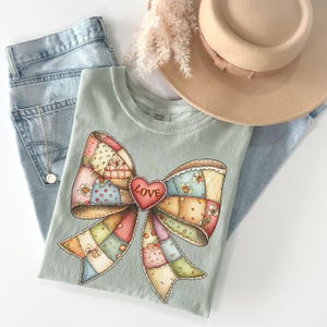 May include: A light green t-shirt featuring a large, colorful bow design with a patchwork pattern and a red heart that says "LOVE". The shirt is paired with light blue jeans and a beige hat.
