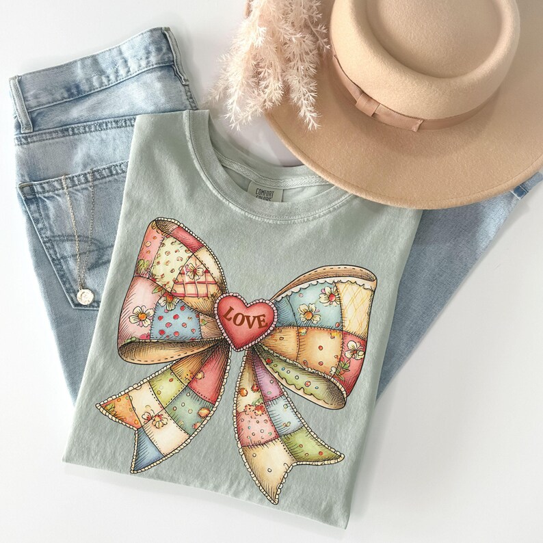 May include: A light green t-shirt featuring a large, colorful bow design with a patchwork pattern and a red heart that says "LOVE". The shirt is paired with light blue jeans and a beige hat.