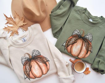 May include: Two sweatshirts with a pumpkin design. One sweatshirt is beige with a black and white lace bow on the pumpkin. The other sweatshirt is olive green with a black and white lace bow on the pumpkin.