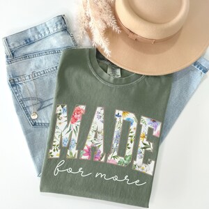 May include: An olive green t-shirt with the word "MADE" in large floral letters and the phrase "for more" in script. The shirt is paired with light wash jeans, a gold necklace, and a tan hat. Dried flowers are also present.