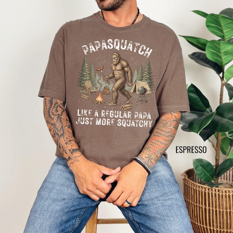 May include: Espresso-coloured t-shirt featuring the text "Papasquatch" and a Bigfoot graphic at a campsite. The graphic includes a campfire, tent, and trees. The text "Like a regular papa, just more squatchy" is also printed on the shirt.
