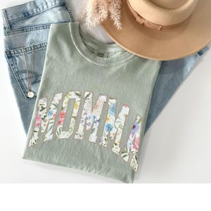 May include: A sage green t-shirt with the word "MOMMA" printed in white with a floral pattern. The shirt is laid out on a light blue denim background.