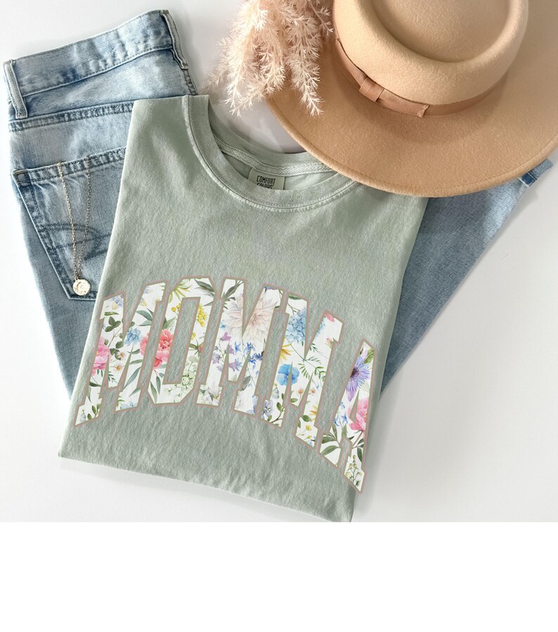 May include: A sage green t-shirt with the word "MOMMA" printed in white with a floral pattern. The shirt is laid out on a light blue denim background.
