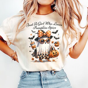 May include: Cream-colored t-shirt with the text "Just A Girl Who Loves Pumpkin Spice." The graphic features a ghost wearing sunglasses, a bow, and holding a pumpkin spice latte and a jack-o'-lantern bag. Black bats and fall leaves surround the ghost.