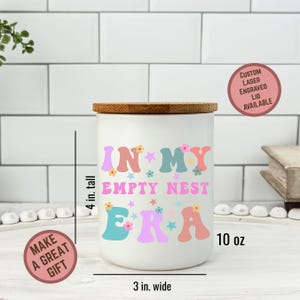 Op de afbeelding: Een witte keramische pot met een houten deksel, met de tekst "IN MY EMPTY NEST ERA" in kleurrijke letters en bloemmotieven. De pot is ongeveer 10 cm hoog en 7,6 cm breed, met een inhoud van 295 ml. Een op maat gemaakte lasergegraveerde deksel is beschikbaar.