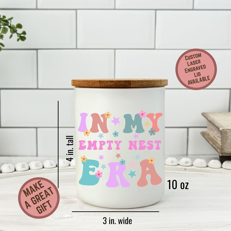 Op de afbeelding: Een witte keramische pot met een houten deksel, met de tekst "IN MY EMPTY NEST ERA" in kleurrijke letters en bloemmotieven. De pot is ongeveer 10 cm hoog en 7,6 cm breed, met een inhoud van 295 ml. Een op maat gemaakte lasergegraveerde deksel is beschikbaar.