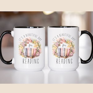May include: Two white ceramic mugs with black handles and interior. Each mug features the text "IT'S A BEAUTIFUL DAY for READING" with a floral wreath and a stack of books. The mugs are designed for book lovers.