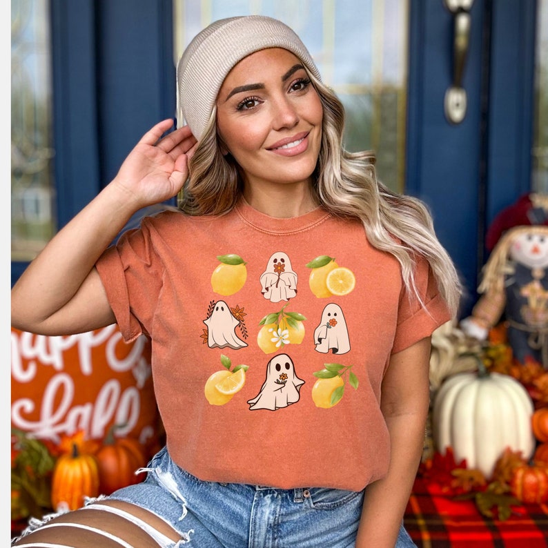 May include: A burnt orange t-shirt with a graphic of ghosts holding lemons and flowers. The ghosts are white and the lemons are yellow. The flowers are orange and white.